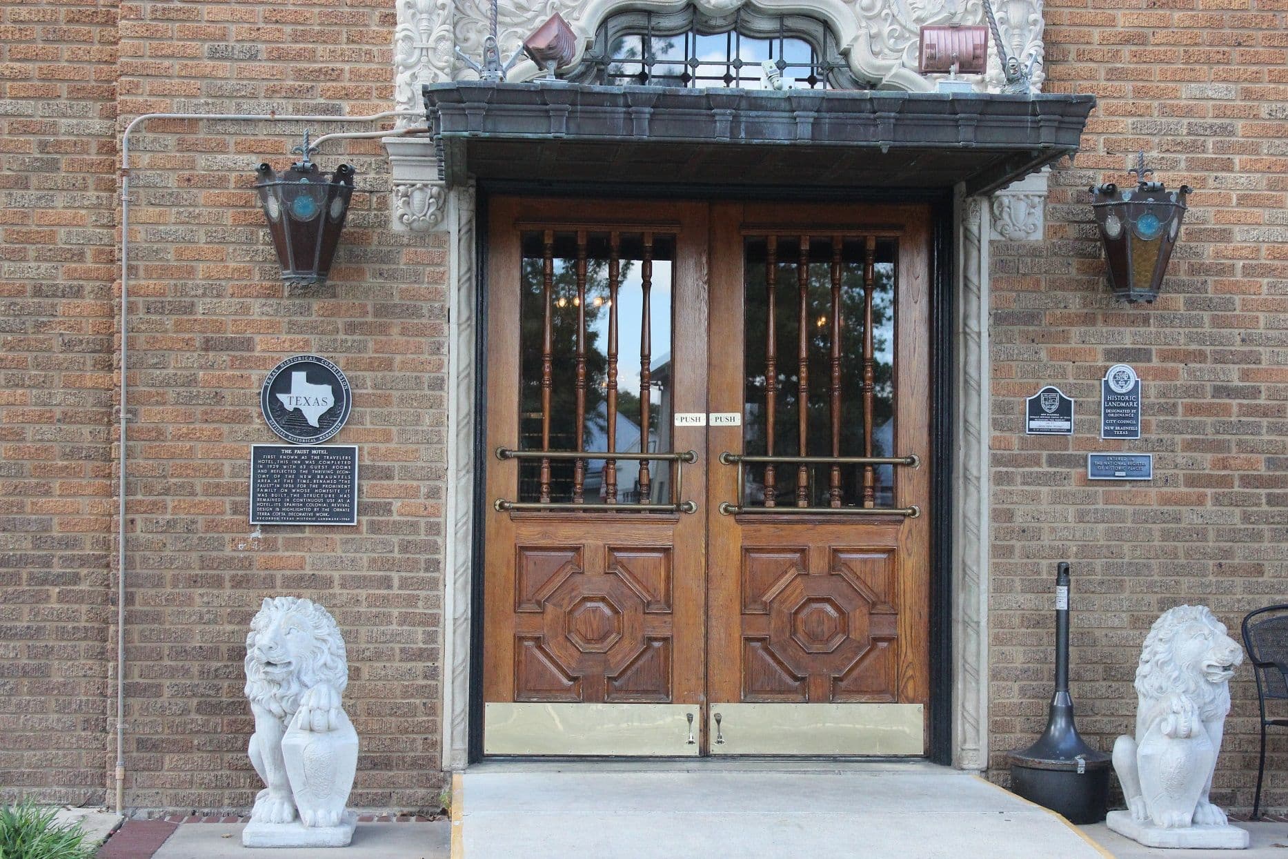 A recessed hotel entrance in a brown and tan brick wall: two heavy panelled oak doors with spindled glazing and brass push bars under a metal canopy, ornate moulded trim above, large wrought-metal lanterns to either side, cast plaques on the brickwork, and a white seated lion statue on each side of the concrete apron.