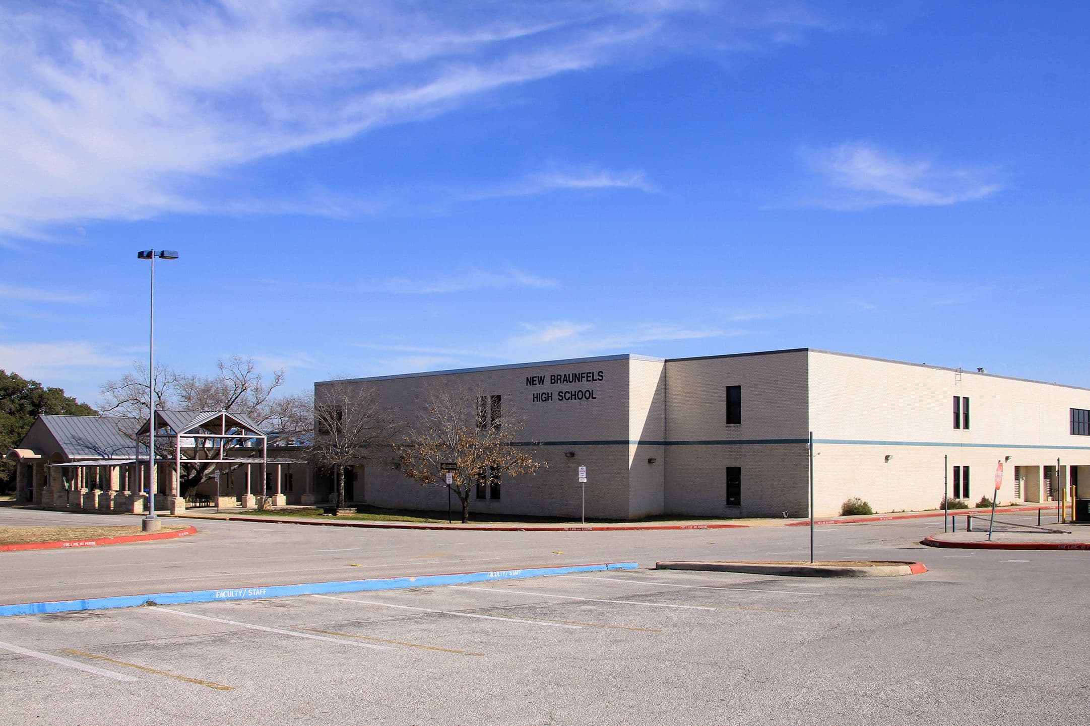 A long two-storey buff brick school building with a grey end wall lettered NEW BRAUNFELS HIGH SCHOOL. A lower limestone wing with a metal gable roof stands to the left, and an empty parking lot with blue-painted kerbs fills the foreground.