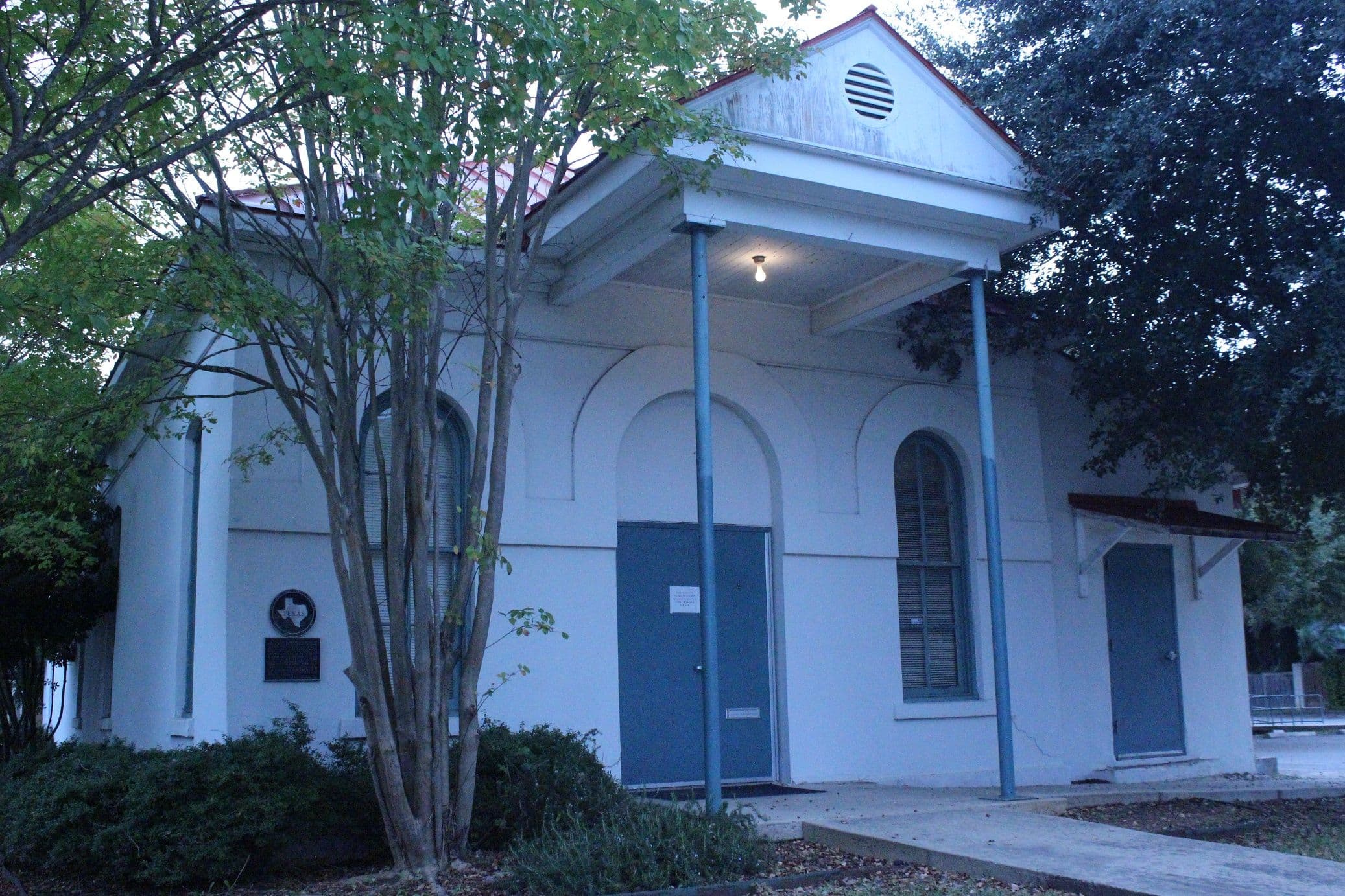 A small single-storey rendered building at dusk, with a triangular pediment over a shallow porch carried on two thin metal posts. Blue-painted double doors sit in a recessed arch, a tall arched louvred window is to the right, and a crepe myrtle stands in front.