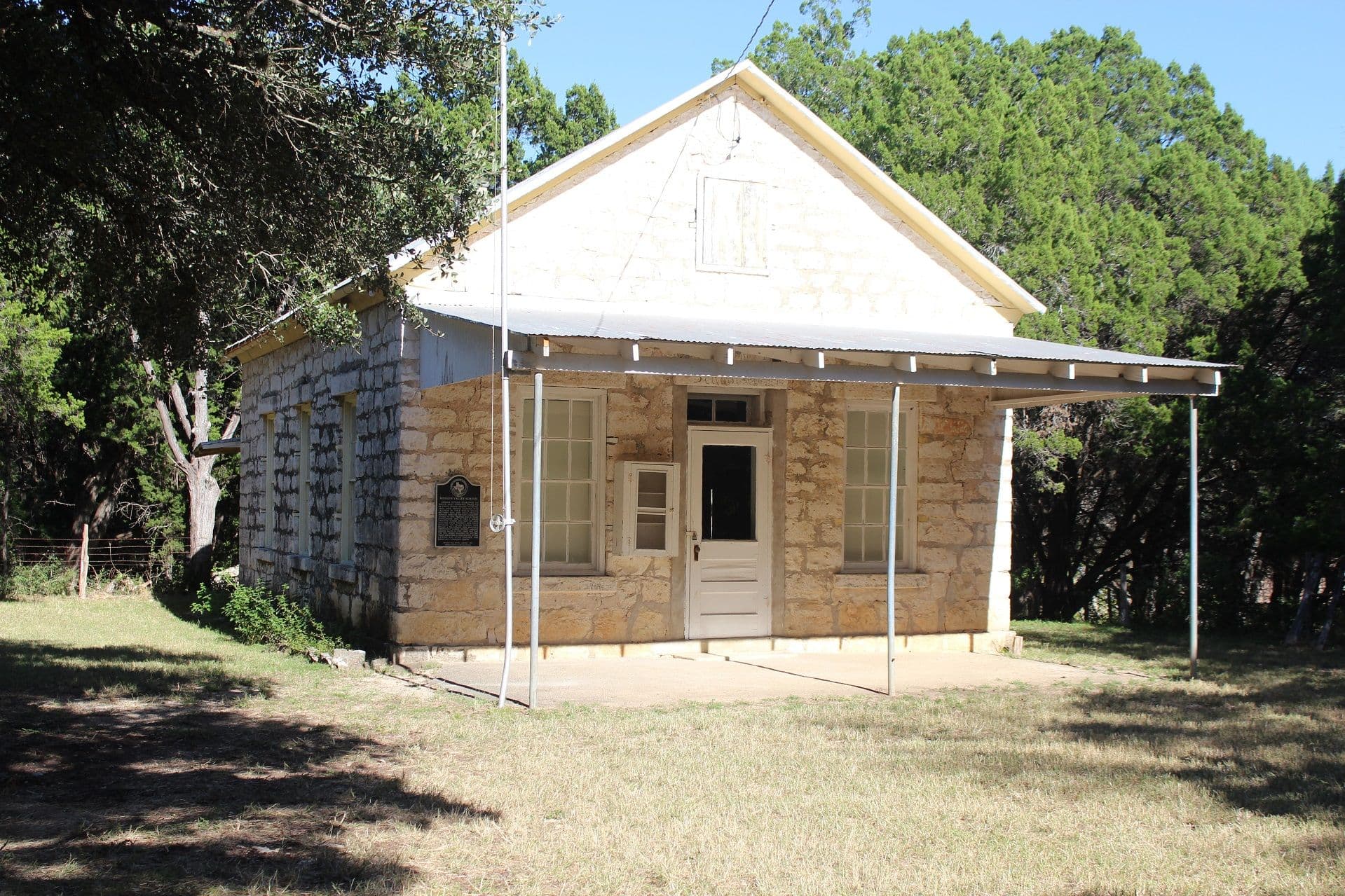 Small single-storey building of buff limestone blocks with a steep gabled metal roof, a corrugated shed porch on thin pipe posts, tall multi-pane windows and a panelled front door, set in mown grass with dense cedar trees behind.