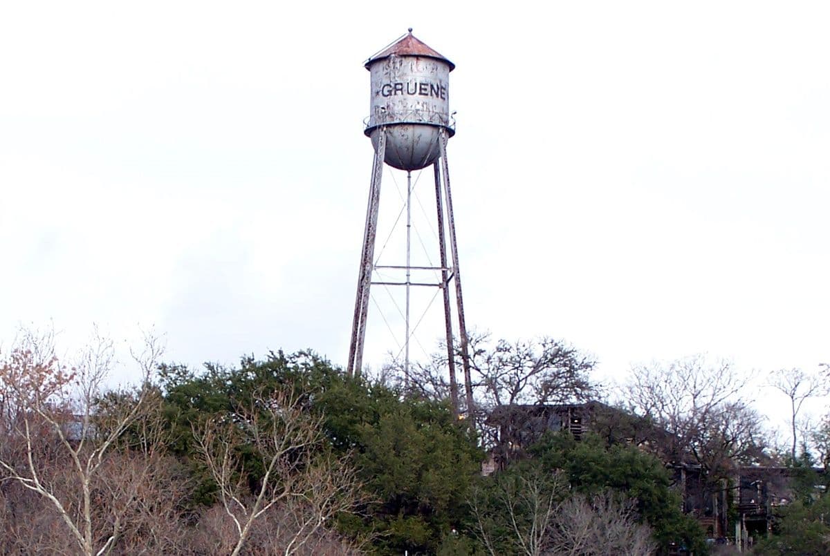 A riveted steel water tower on four lattice-braced legs, its cylindrical tank painted with the name "GRUENE" and streaked with rust under a conical roof, standing above a canopy of bare and evergreen trees against a white sky.