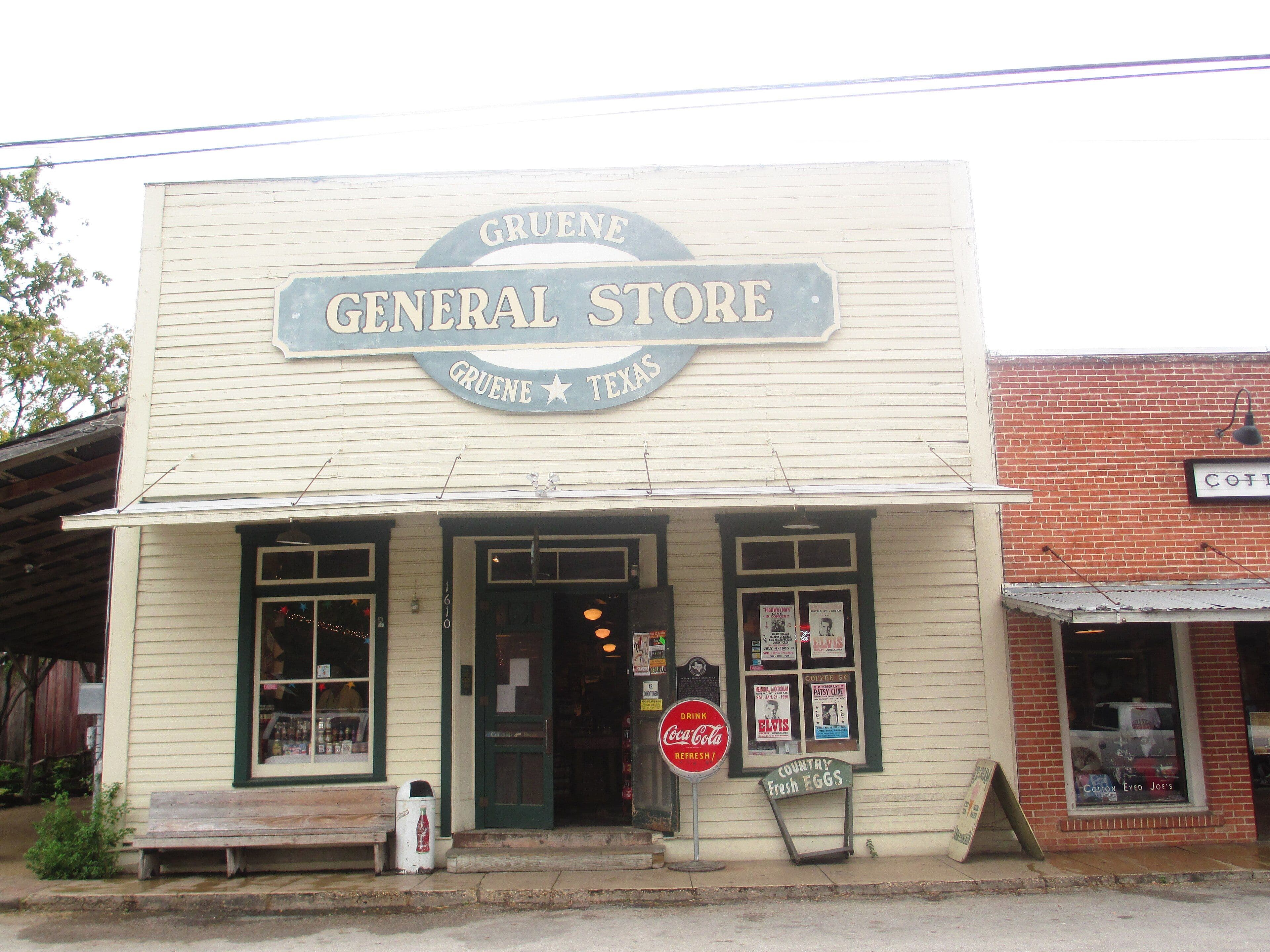 A cream-painted wooden storefront with a tall false front carrying a large oval sign reading Gruene General Store, a narrow metal awning over an open screen door, display windows with posters, and a round Coca-Cola sign on a post outside.