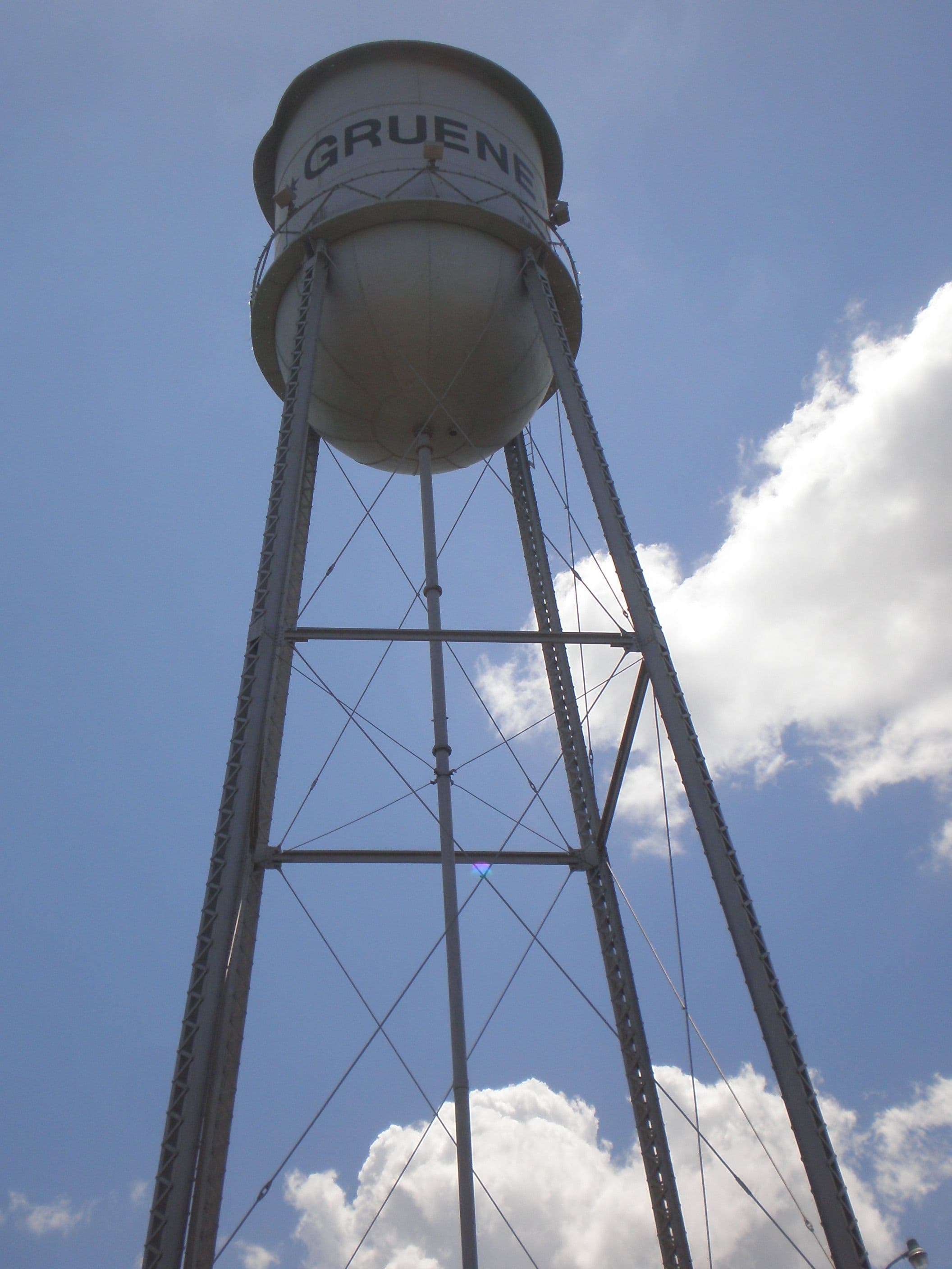 Looking steeply up at a white elevated steel water tank lettered GRUENE, carried on four splayed lattice legs with diagonal bracing, against a blue sky with white cumulus clouds.