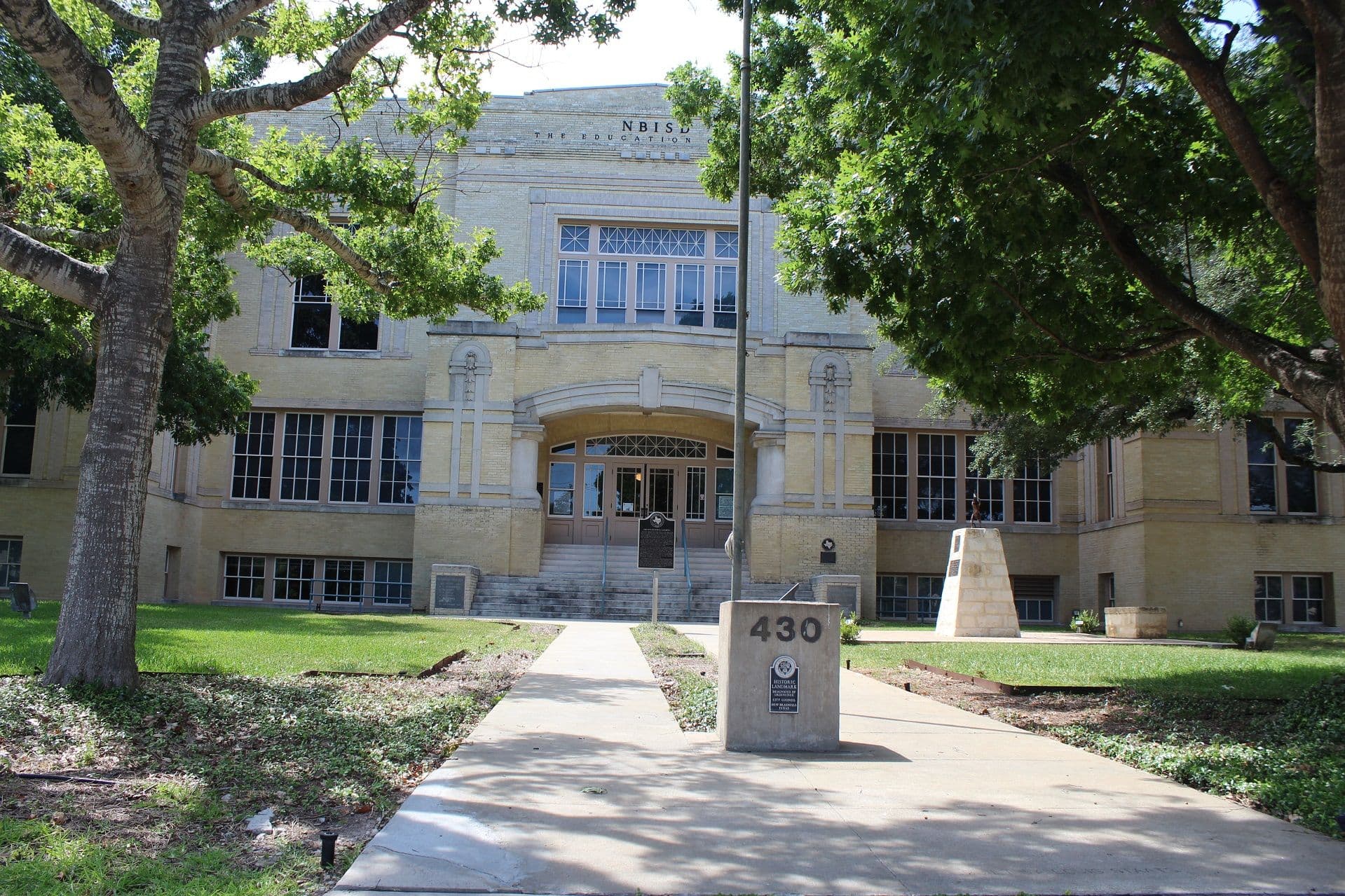 A large symmetrical two-storey building of pale buff brick with a raised arched central entrance up a broad flight of concrete steps, tall multi-pane windows in both wings, NBISD painted high on the parapet, a concrete address pier on the front walk and mature trees framing the view.