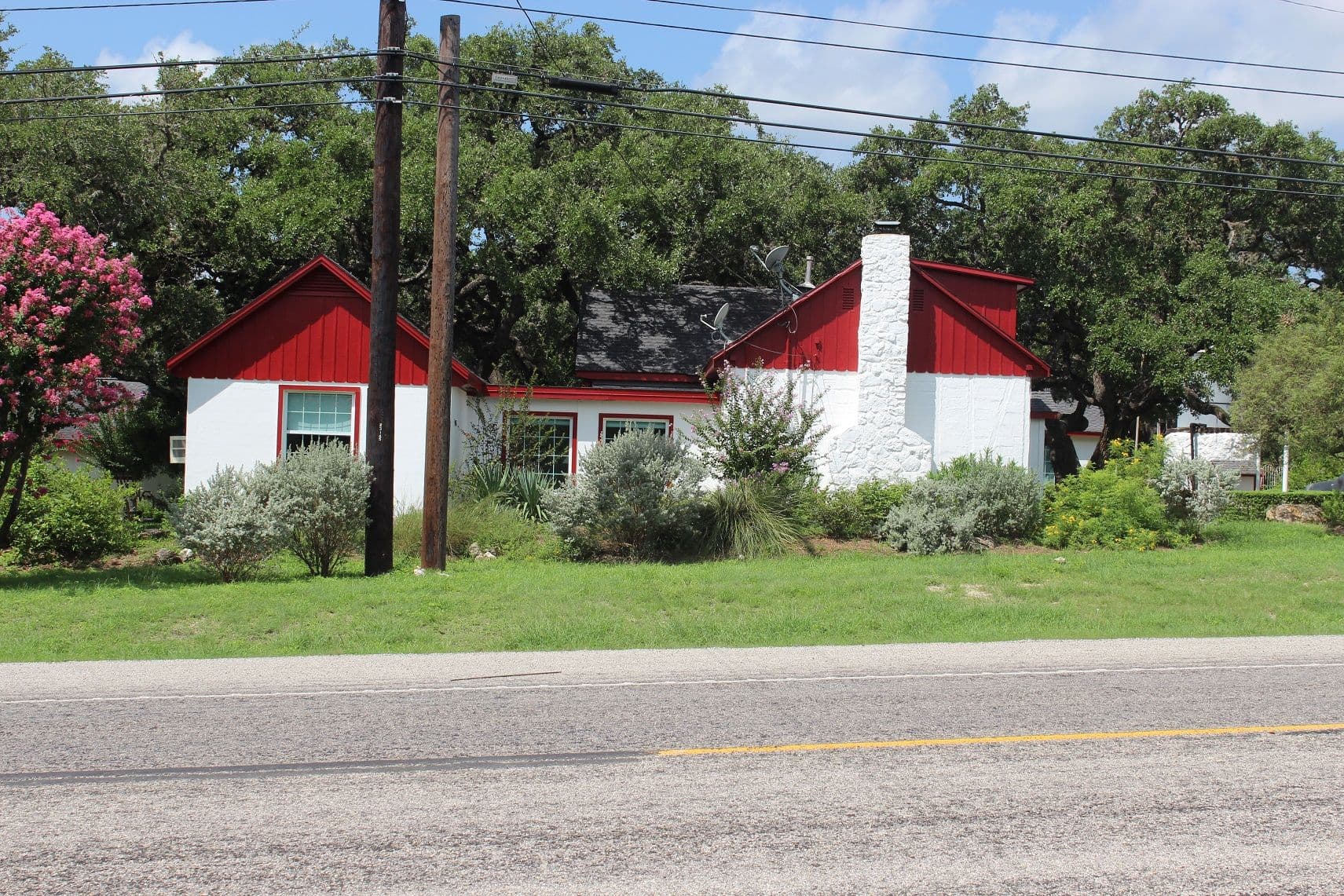 A low white building with red-painted gable ends and window trim stands behind shrub planting on a grassy rise across a two-lane road. A tall whitewashed stone chimney rises through the roof and large live oaks stand behind.