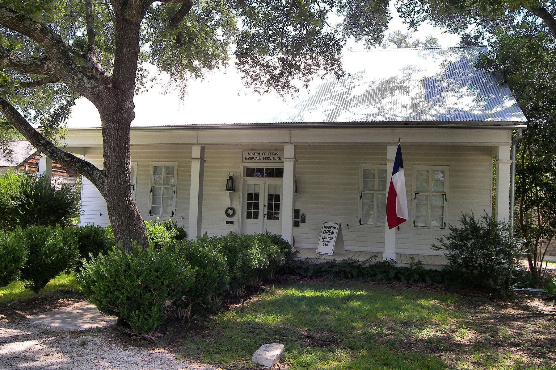 A single-storey white clapboard house with a corrugated metal roof and a deep full-width porch. Lettering on the porch frieze reads Museum of Texas Handmade Furniture; a Texas flag hangs from a porch post and clipped hedges line the front.