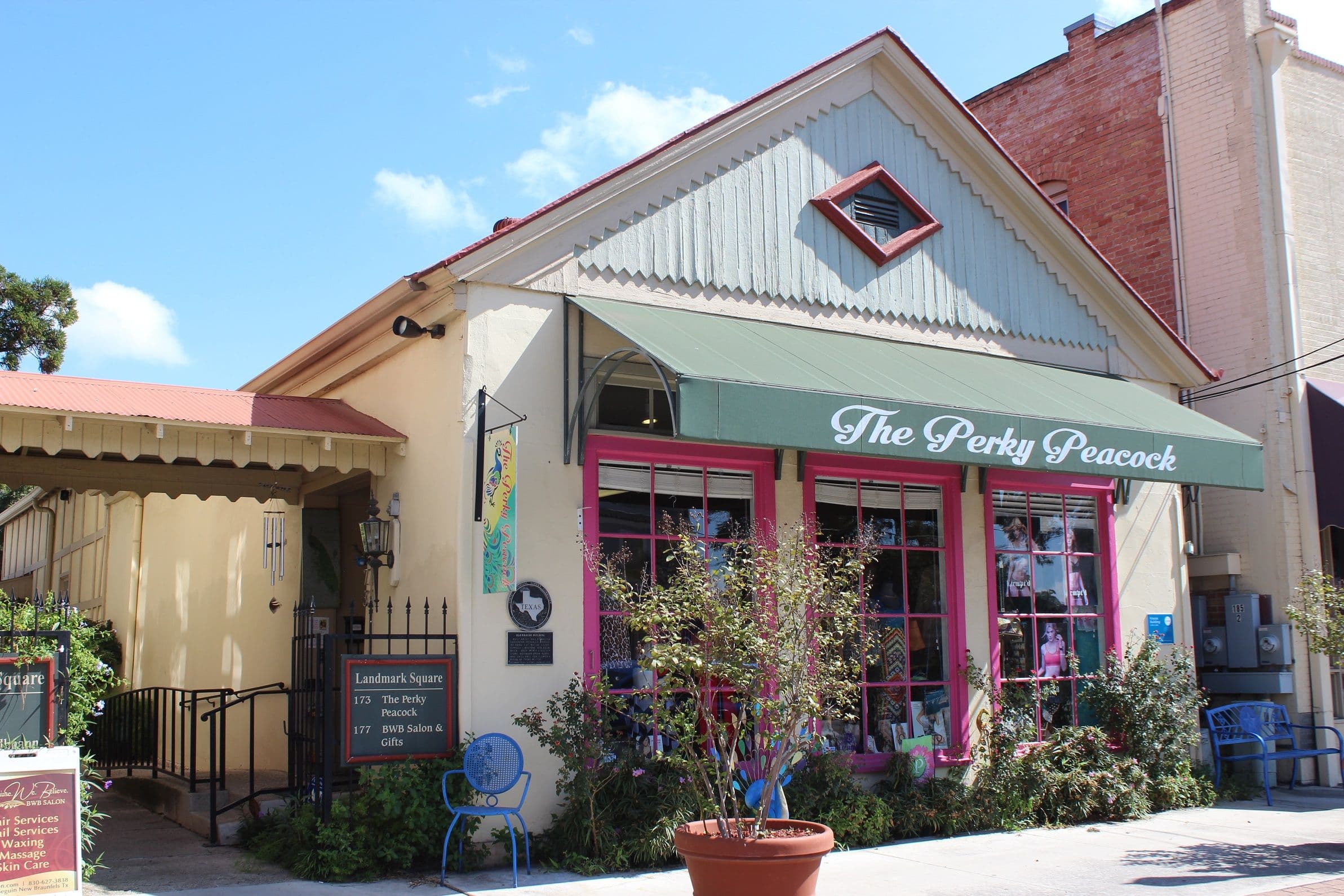 A small cream stuccoed building with a steep board-and-batten gable and a red metal roof. A sage-green awning lettered The Perky Peacock shelters a shopfront with magenta window frames, and a painted directory board listing two tenants stands at the left beside an iron gate. A young tree in a terracotta pot sits on the sidewalk.
