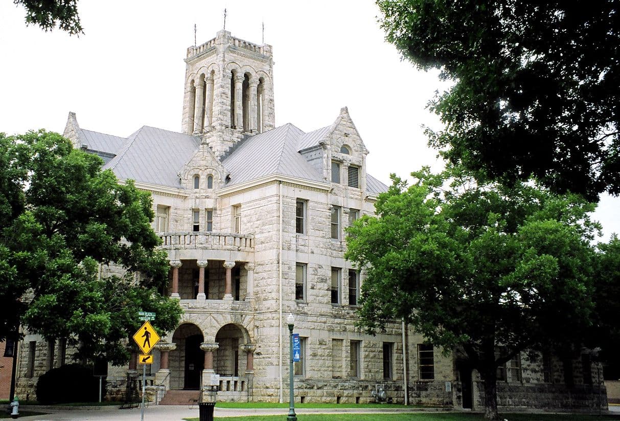 A three-storey rock-faced limestone building with a steep grey hipped roof, gabled dormers and a square arcaded tower with a crenellated top. A broad round-arched entrance loggia sits at the centre with a stone balcony above. Mature oaks frame the view and a yellow pedestrian crossing sign stands on the sidewalk.