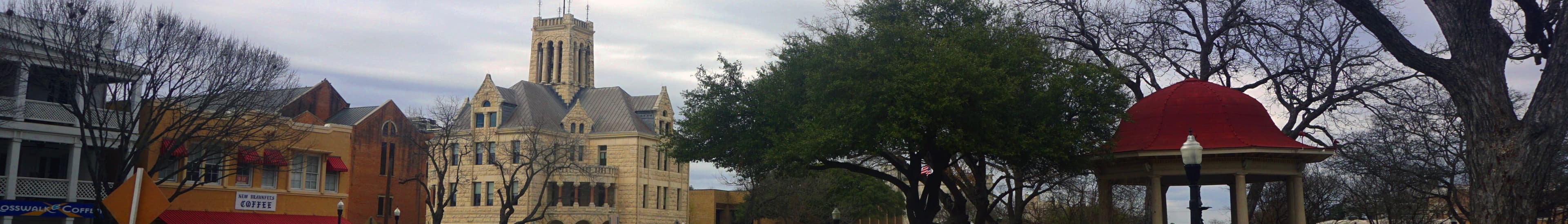A very wide, short panorama of a town plaza under grey cloud: low storefronts with red awnings at the left, a three-storey rusticated stone building with steep gables and a square tower at the centre, large trees, and an open pavilion with a red dome at the right.