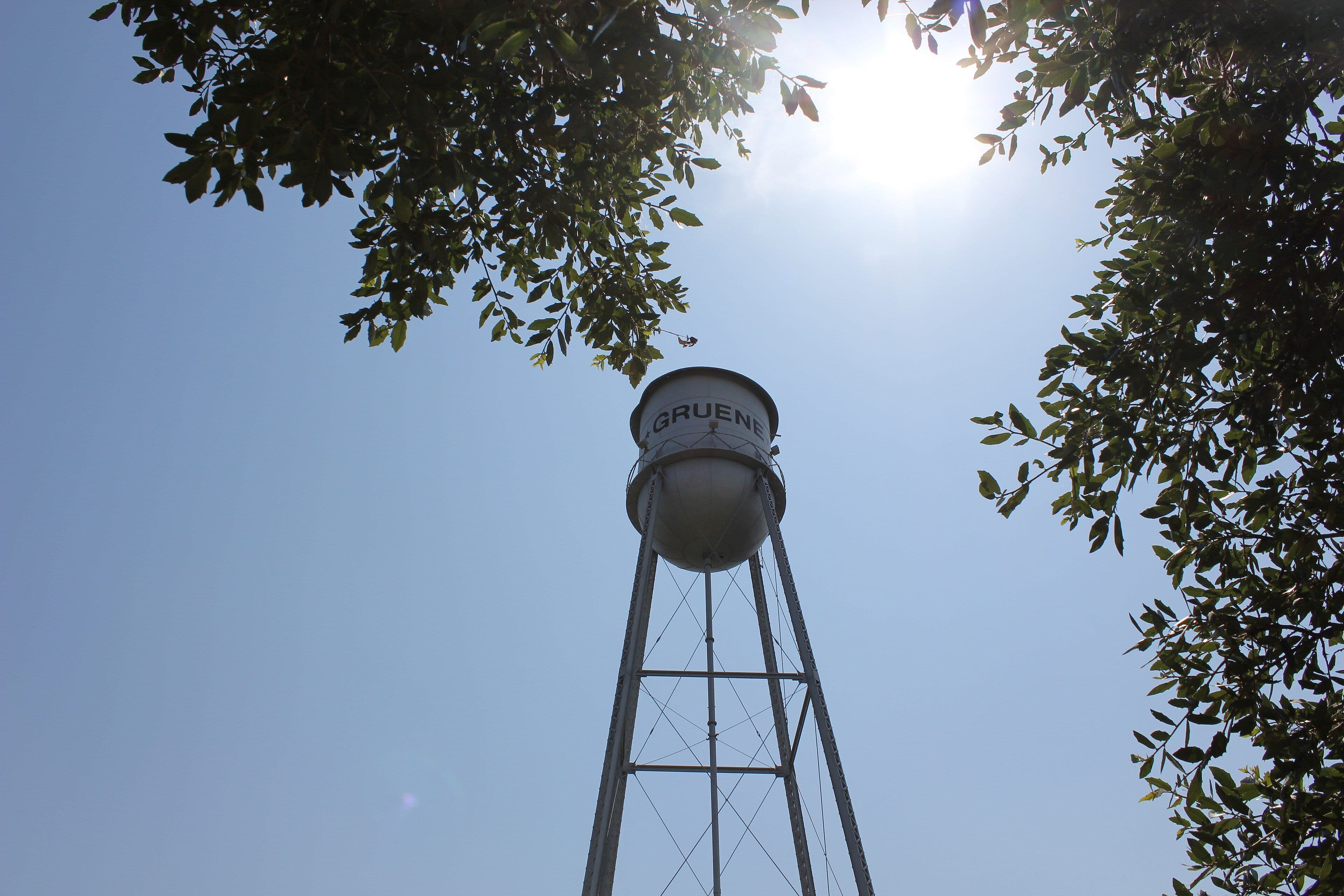 Looking steeply upward at a silver-white steel water tower lettered GRUENE around its tank, standing on four tall lattice legs against a pale blue sky, with dark oak leaves framing the top corners and the sun flaring behind them.