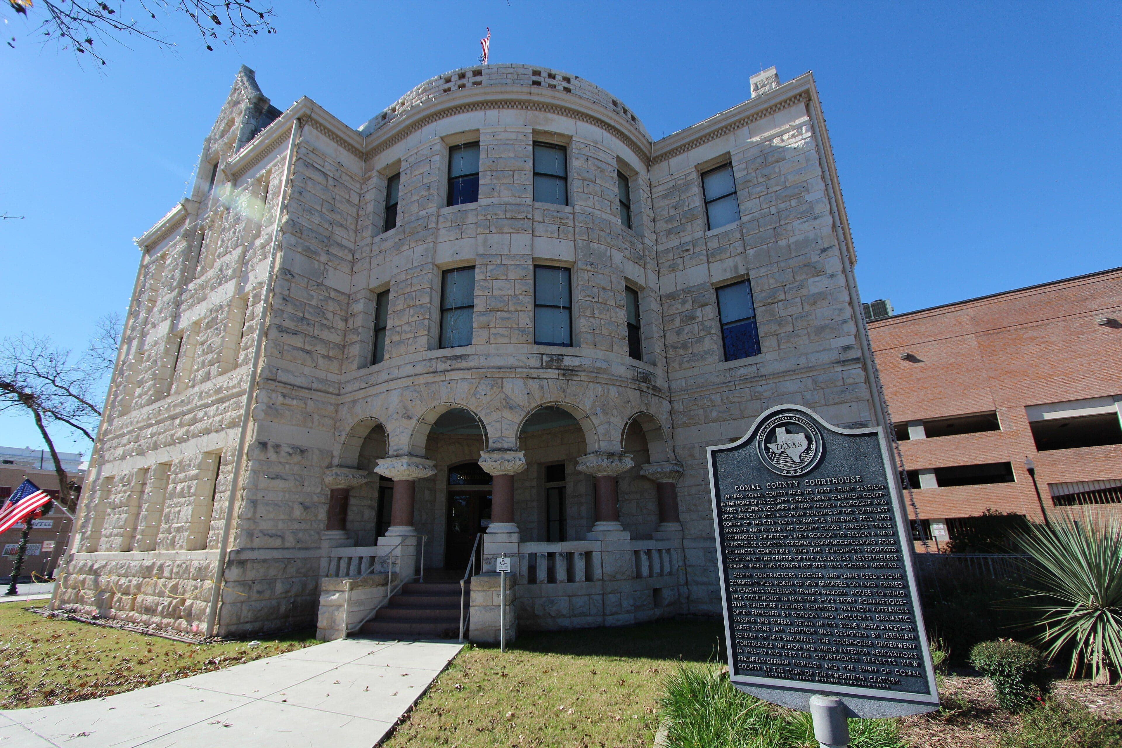 A three-storey rusticated limestone courthouse photographed from below against a clear blue sky, with a rounded central pavilion, an arcaded ground-floor entrance on short columns, a flag on the roof and a dark metal historical marker on a post at right.
