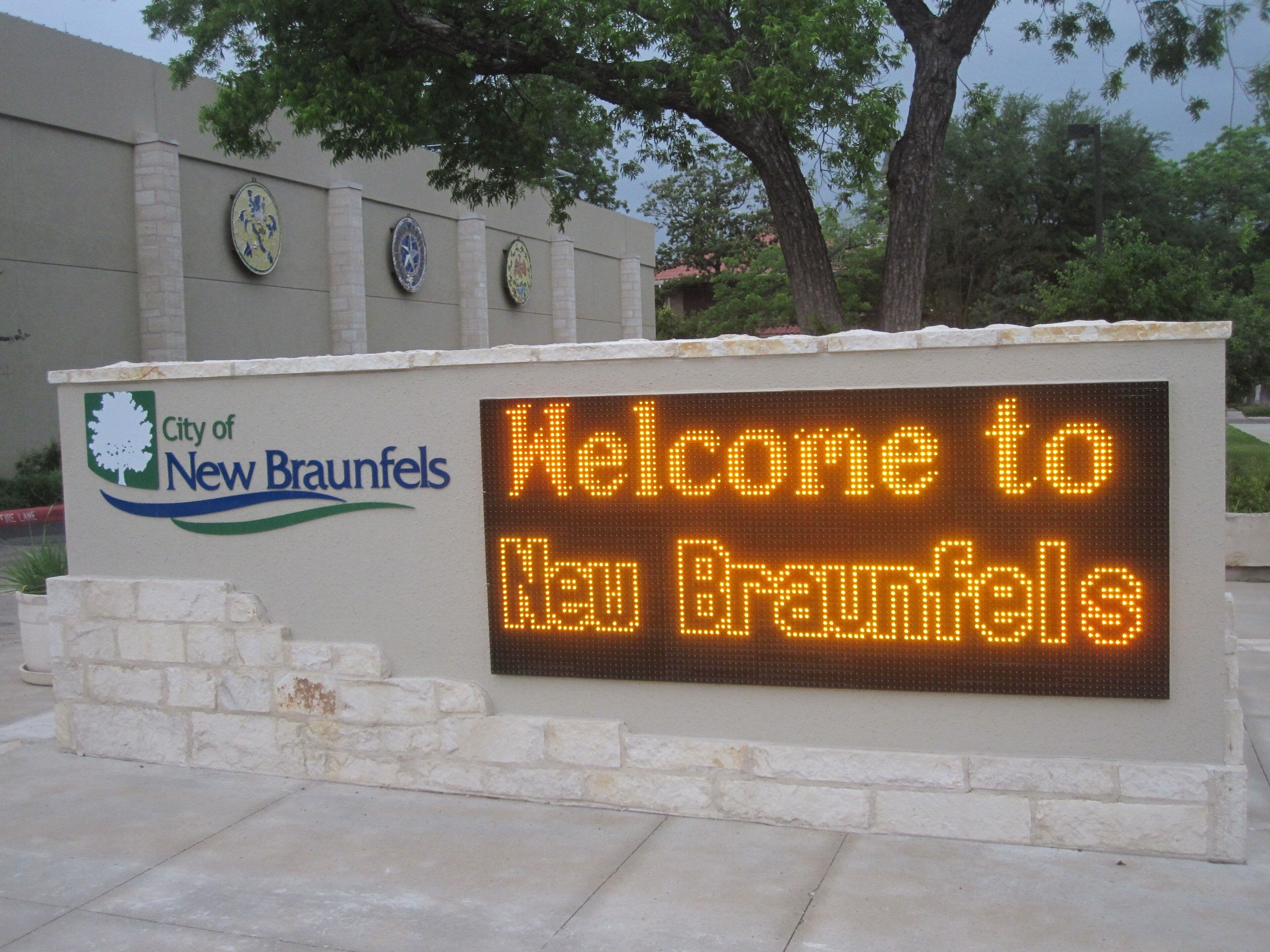 A low rendered wall capped with rough limestone carries the City of New Braunfels logo at the left and a large amber LED board reading “Welcome to New Braunfels” at the right. A rendered building with three circular ceramic medallions stands behind under a grey sky.