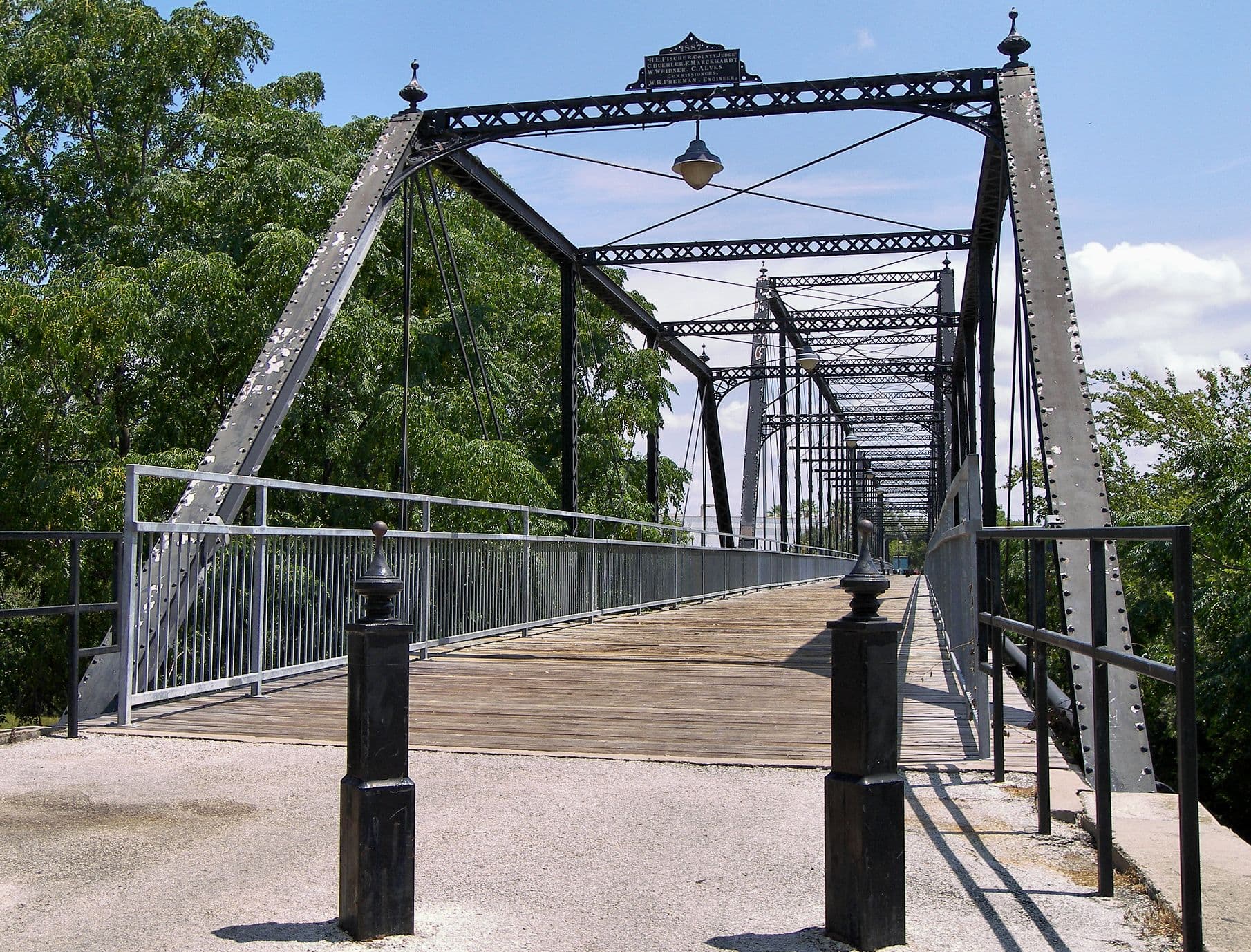 A metal truss bridge seen head-on from its approach. Two inclined riveted members rise to an arched lattice top chord with urn-shaped finials, and a cast plate and a hanging lamp are fixed beneath it. Truss panels recede in perspective down the bridge. The deck is laid with transverse timber planks between pipe railings, and two square metal posts stand on the gravel approach in the foreground. Green trees crowd both sides.
