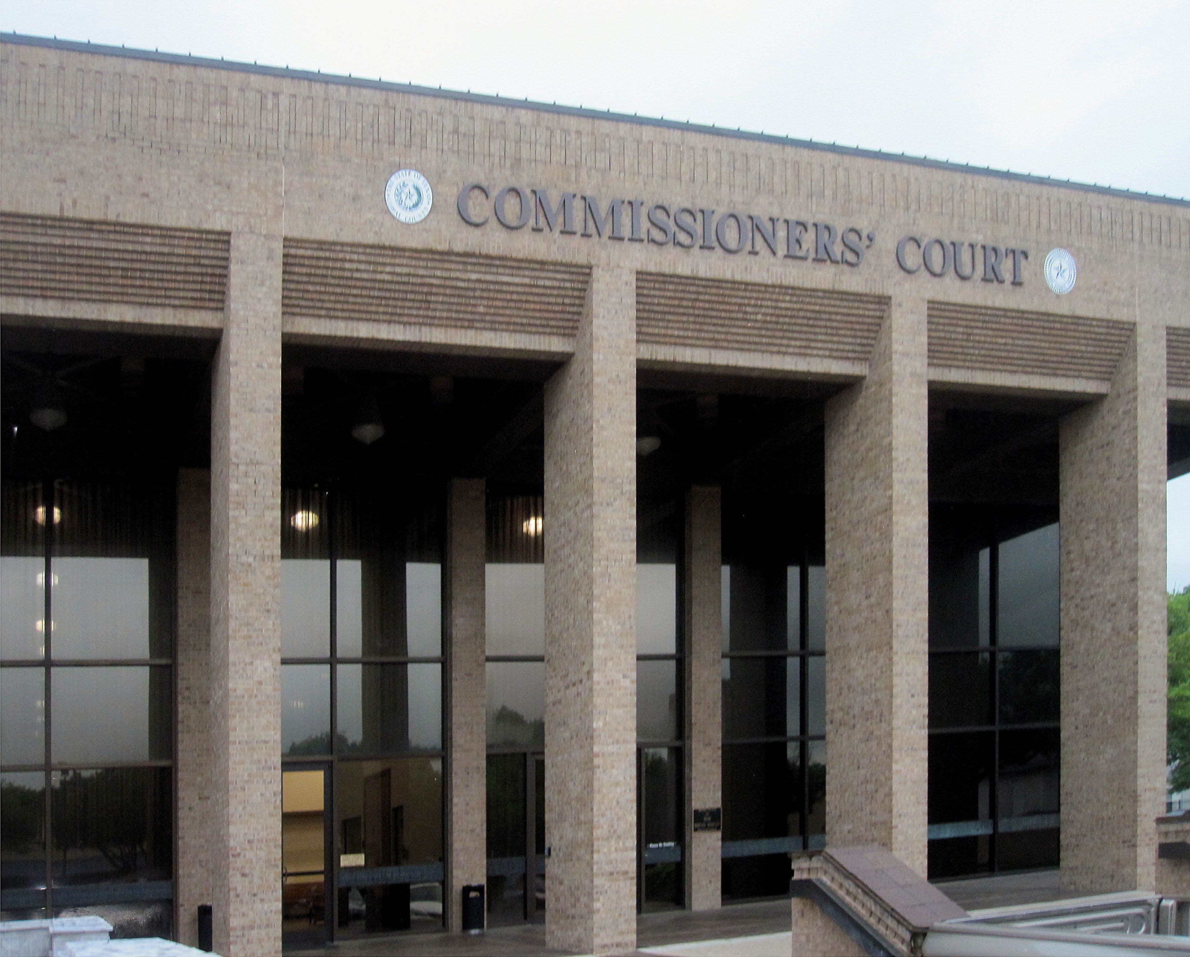The upper front of a modern buff brick public building. Tall square brick piers form a deep shaded porch in front of a glass wall, and raised metal letters spelling COMMISSIONERS' COURT run along the brickwork above, between two round metal seals.